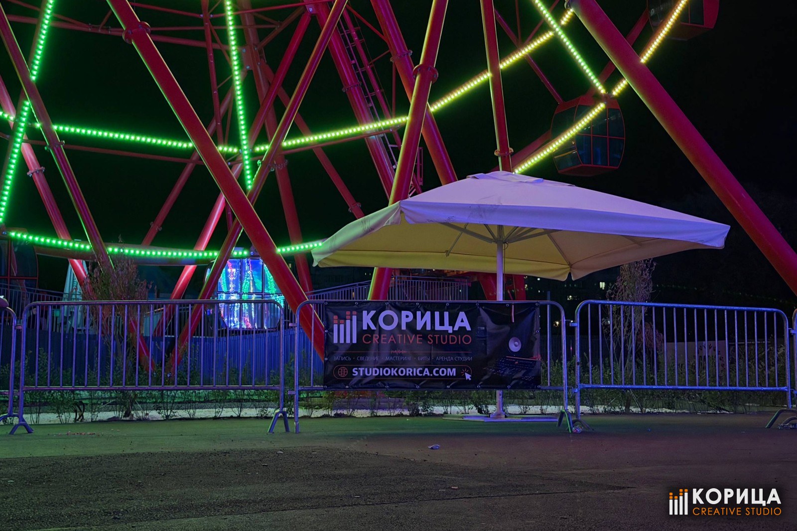Баннер студии «Корица» на ограждении площадки вечером, подсветка аттракционов